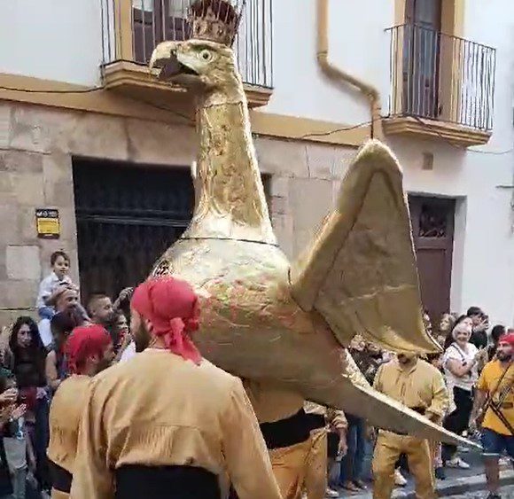 Figura tradicional de un águila dorada gigante (l'Àliga) llevada por personas en la calle durante una fiesta popular rodeada de gente.