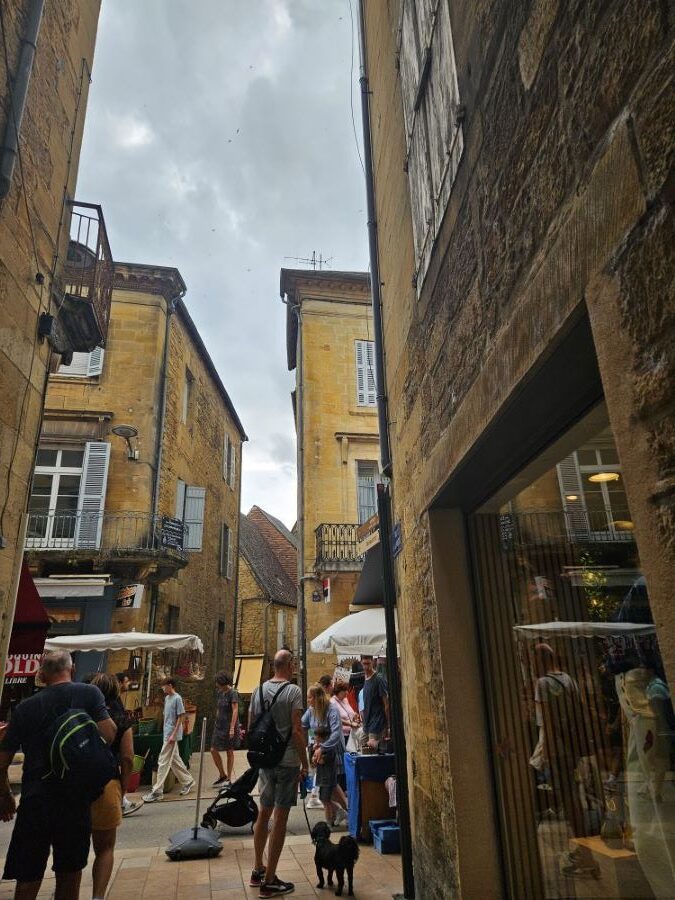 Calle estrecha medieval en Sarlat con edificios de piedra dorada bajo un cielo nublado, con gente paseando entre puestos de mercado sombreados.