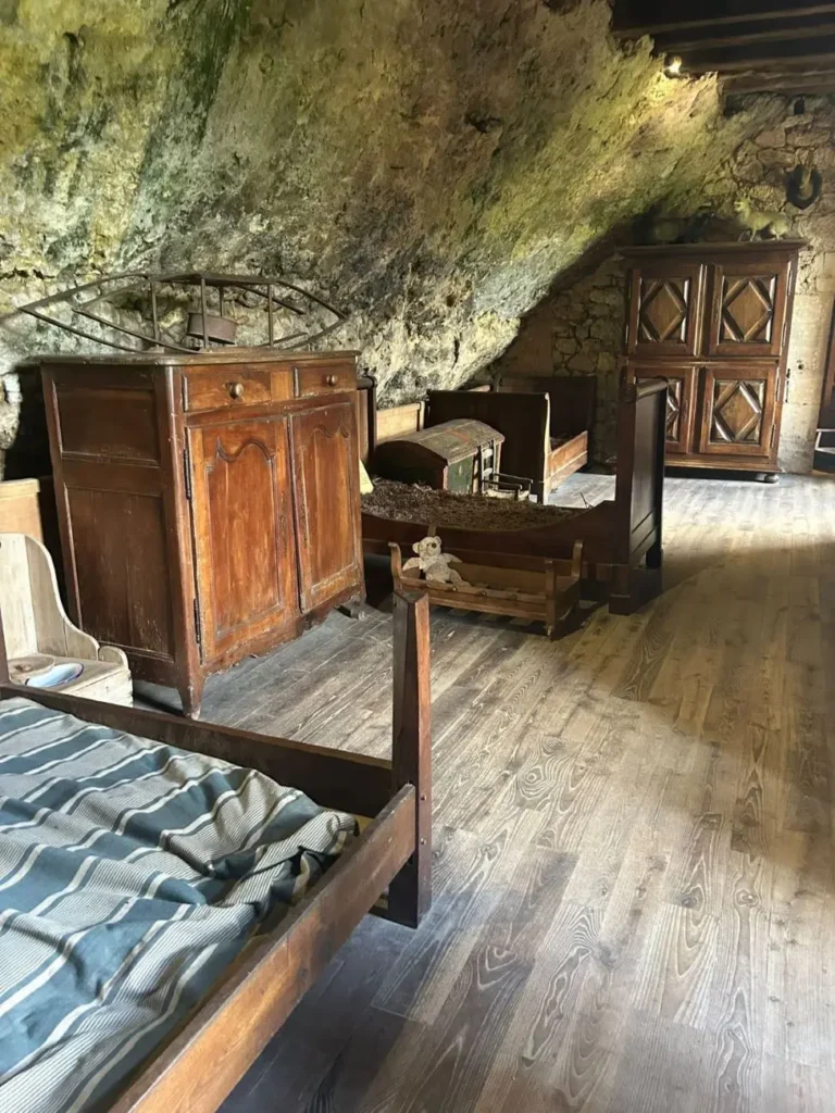 Interior de un dormitorio medieval en la Maison Forte de Reignac, con una cama grande de madera y muebles de época bajo un techo de roca natural irregular.