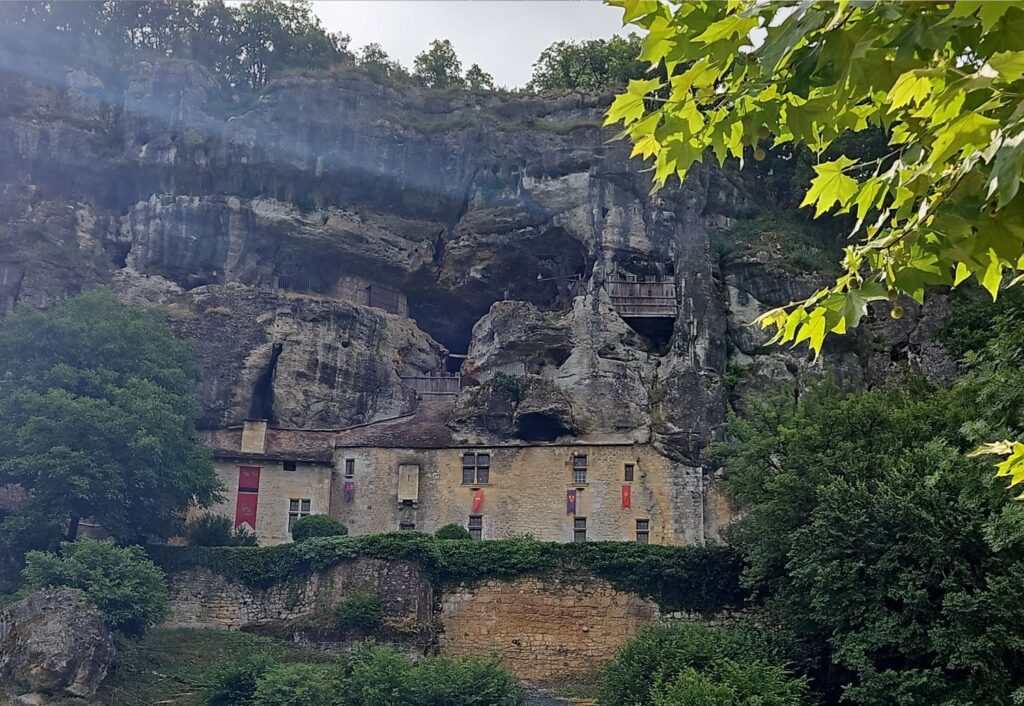 Vista completa del castillo troglodita Maison Forte de Reignac integrado en un enorme acantilado de piedra, mostrando estructuras de madera en las cavidades superiores.