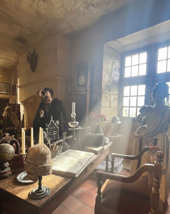 Espectaculares rayos de luz solar entrando por una ventana y cruzando un estudio con un escritorio, libros antiguos y un maniquí en la Maison Forte de Reignac.