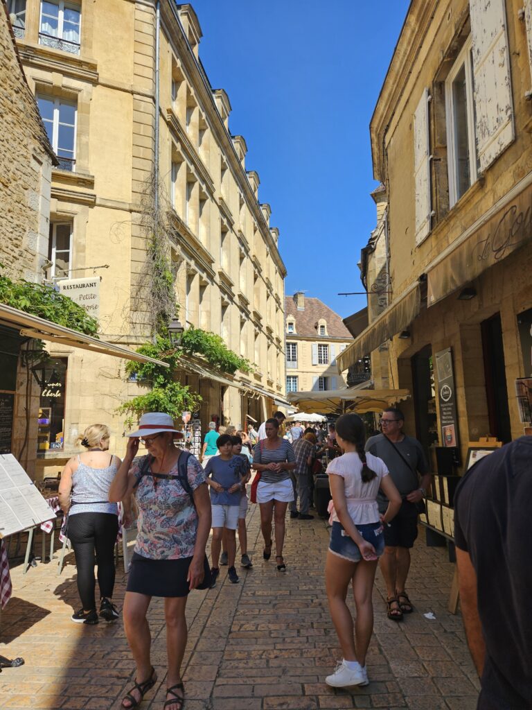 Callejón medieval de Sarlat durante el mercado del miércoles, con edificios de piedra caliza bajo un cielo azul despejado y turistas paseando.