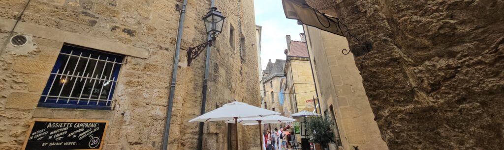 Callejón medieval en Sarlat-la-Canéda con muros de piedra ocre, sombrillas blancas de mercado y pizarra de menú tradicional con "Assiette Campagne
