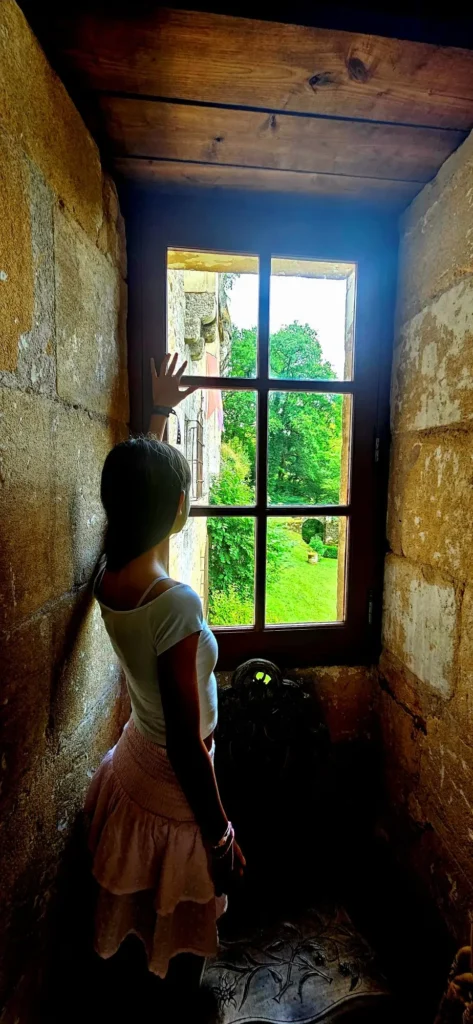 Una mujer mira por una gran ventana hacia el valle soleado desde el interior oscuro y de piedra de la Maison Forte de Reignac.