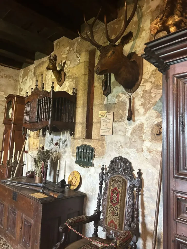 Estancia rústica en la Maison Forte de Reignac con paredes de piedra vista, armarios antiguos de madera, una silla decorada y una cabeza de ciervo montada en la pared.