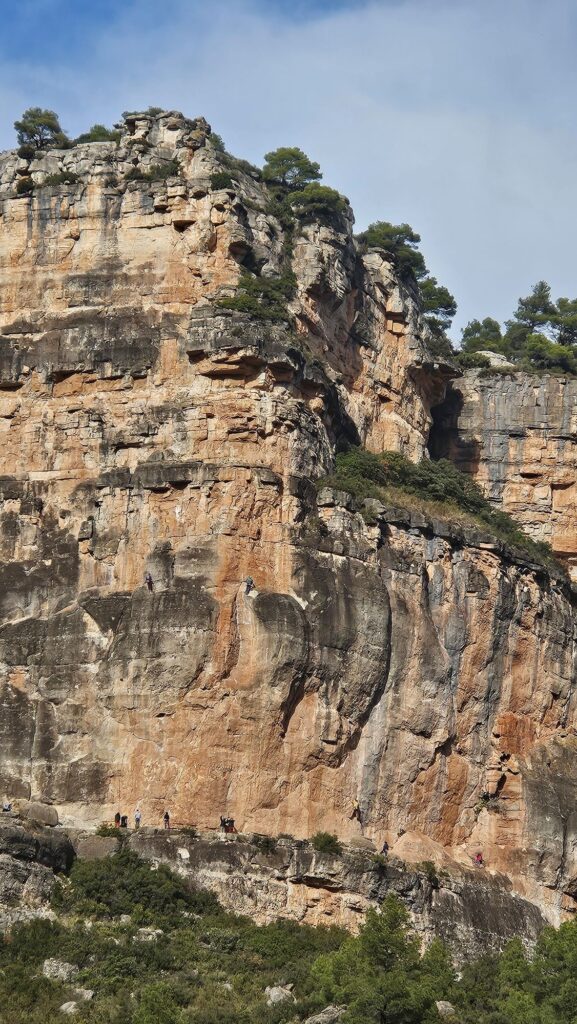 Vista panorámica de los imponentes muros de roca caliza en Siurana con pequeños escaladores ascendiendo, destacando la magnitud del paisaje.