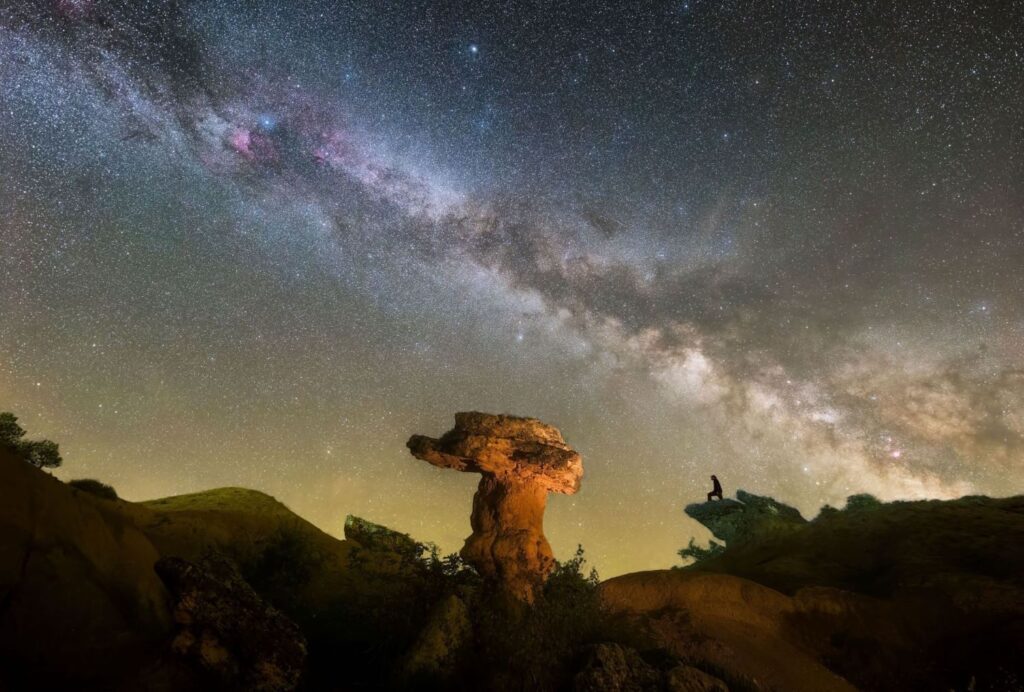 Espectacular fotografía nocturna de la Vía Láctea brillando sobre la formación rocosa conocida como la Roca del Bolet, en la zona de Figuerola de Orcau, cerca del Montsec. Se ve la silueta de una persona observando el cielo, destacando la inmensidad del universo.