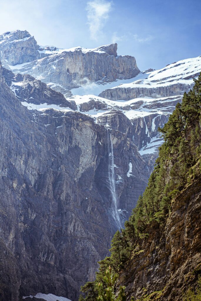 Vista vertical del Circo de Gavarnie en los Pirineos franceses. Se aprecia la inmensa pared rocosa gris con restos de nieve y una larga cascada cayendo hacia el valle, con pinos verdes en primer plano a la derecha.