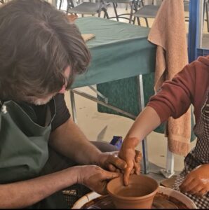 Primer plano de las manos de un alfarero adulto guiando las de un aprendiz en un torno de arcilla. Ilustra la transmisión de la artesanía tradicional.