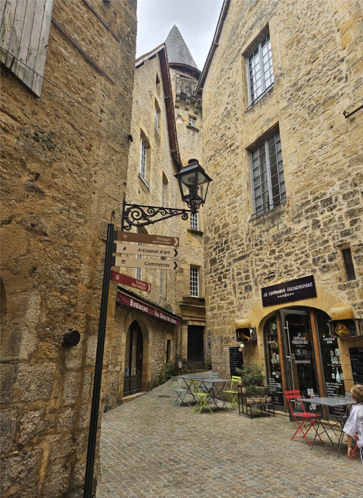 Callejón empedrado en el casco antiguo de Sarlat-la-Canéda con arquitectura de piedra y carteles señalizando talleres de artesanía y arte.