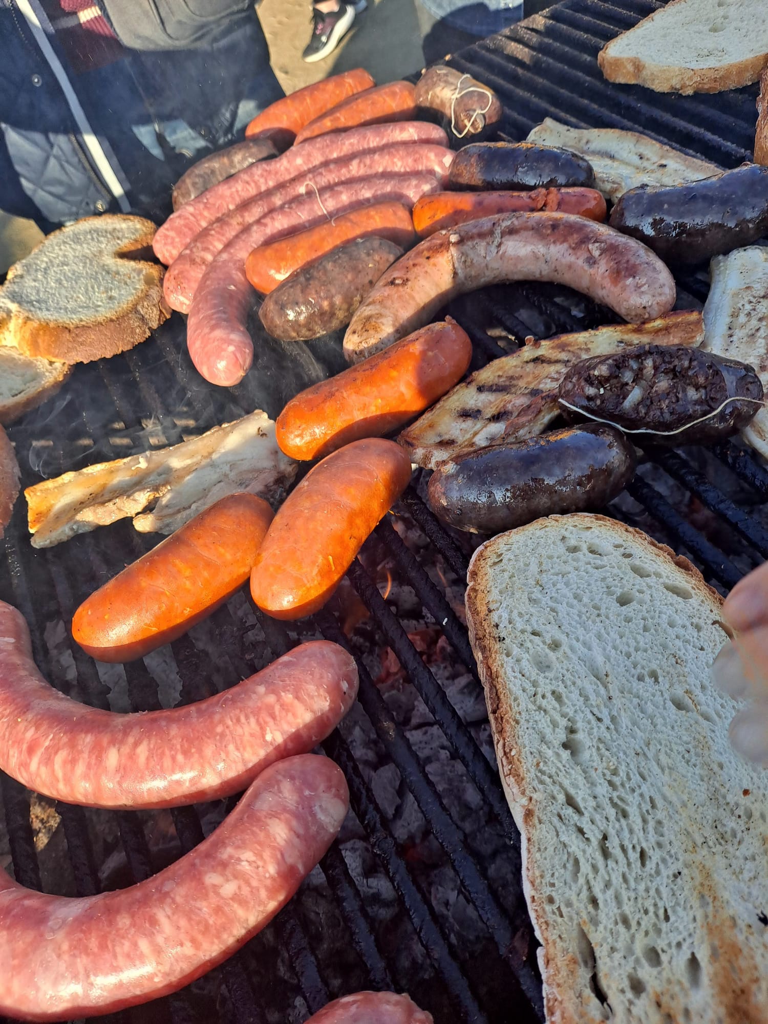 Primer plano de una barbacoa llena de butifarra, morcilla, panceta y pan tostándose, mostrando la variedad de embutidos típicos de un esmorzar popular en Cataluña.