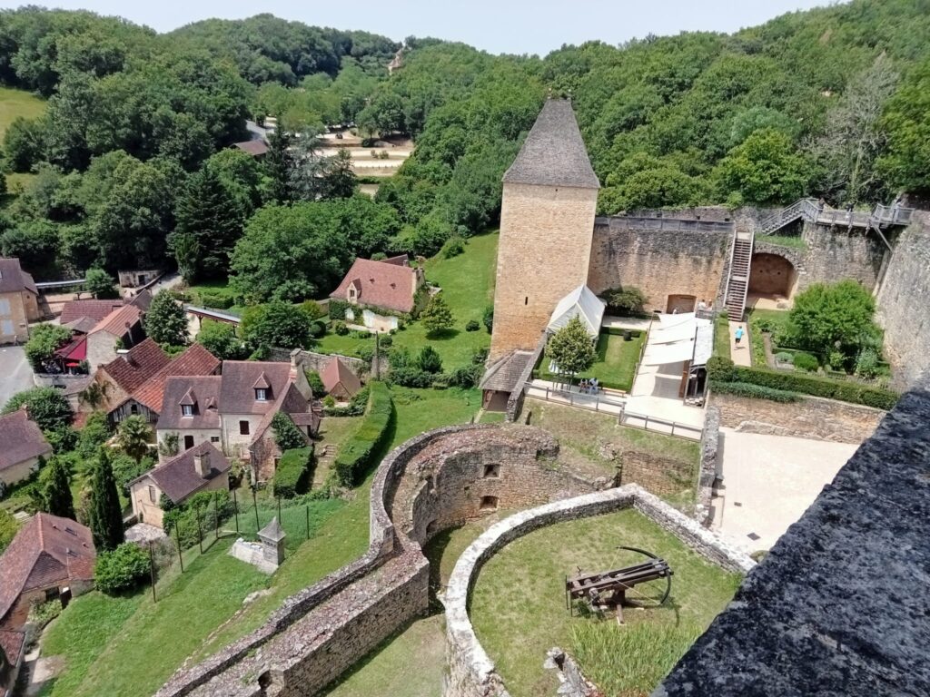 Vista aérea desde las murallas del Castillo de Castelnaud-la-Chapelle en el Périgord. Se observa una catapulta (trebuchet) en el patio de armas, la torre del homenaje cuadrada y el pueblo medieval con tejados rojizos rodeado de bosque.