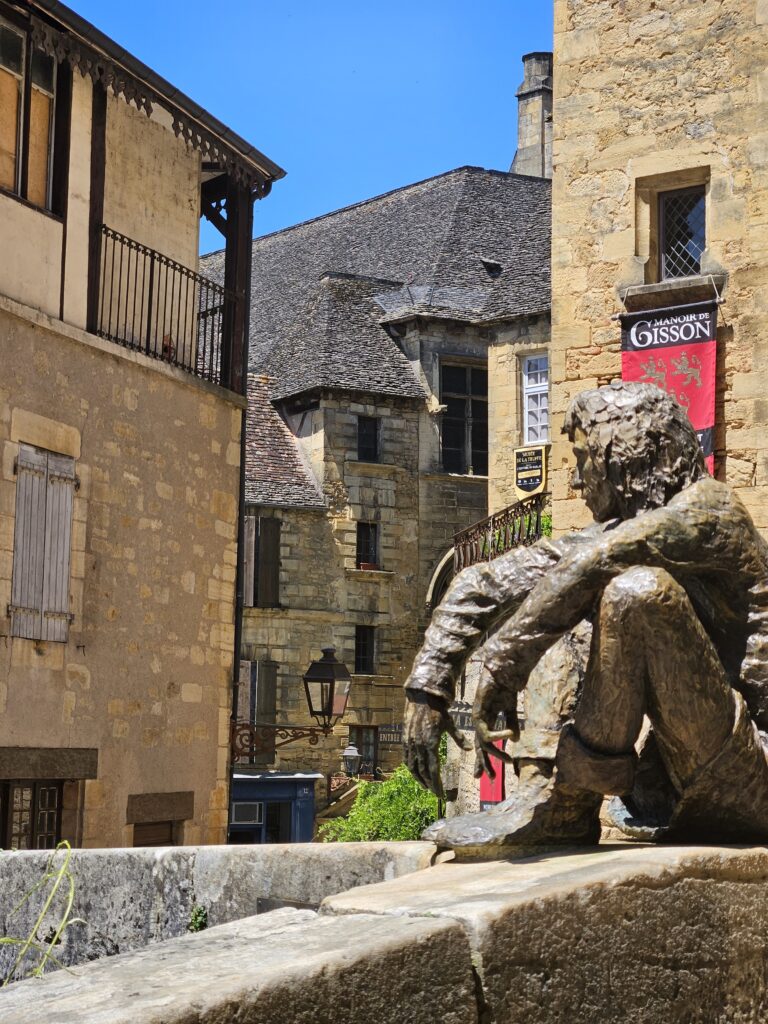 Estatua de bronce de 'Le Badaud' (El Mirón) sentada en un muro de piedra en el casco histórico medieval de Sarlat-la-Canéda, Francia.
