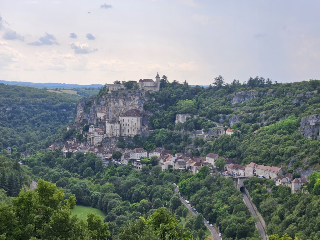 Vista panorámica aérea de un pueblo medieval encaramado en un acantilado y extendiéndose por el valle, rodeado de densa vegetación y una carretera. Simboliza las rutas de leyenda y la inmersión cultural.