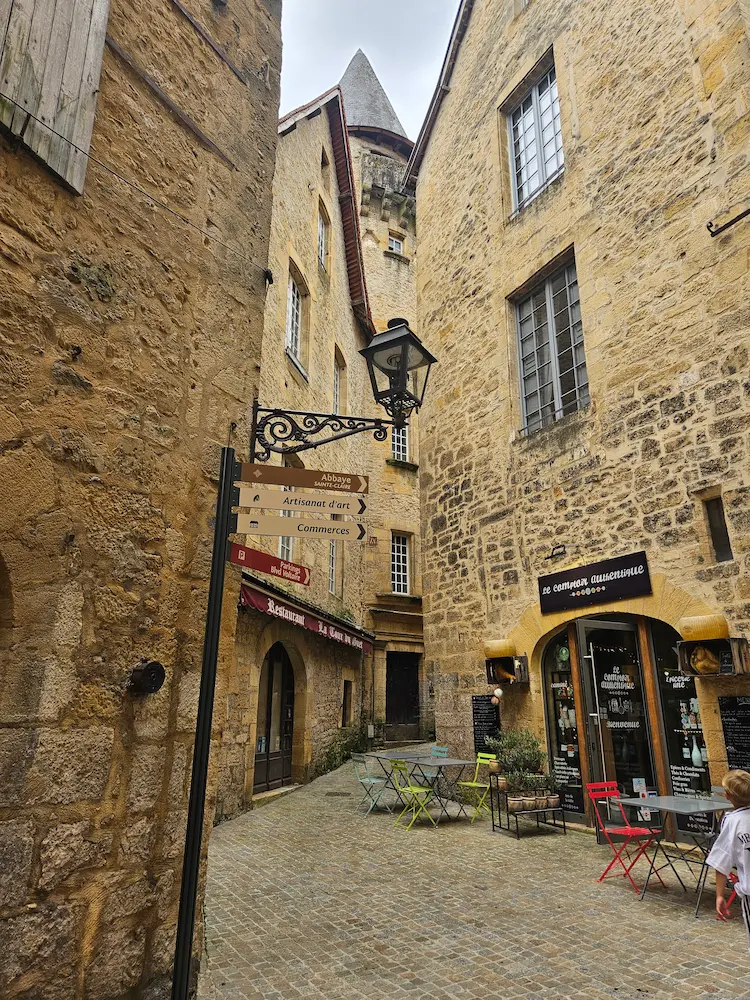 Callejón empedrado en el casco antiguo de Sarlat-la-Canéda con arquitectura de piedra y carteles señalizando talleres de artesanía y arte.