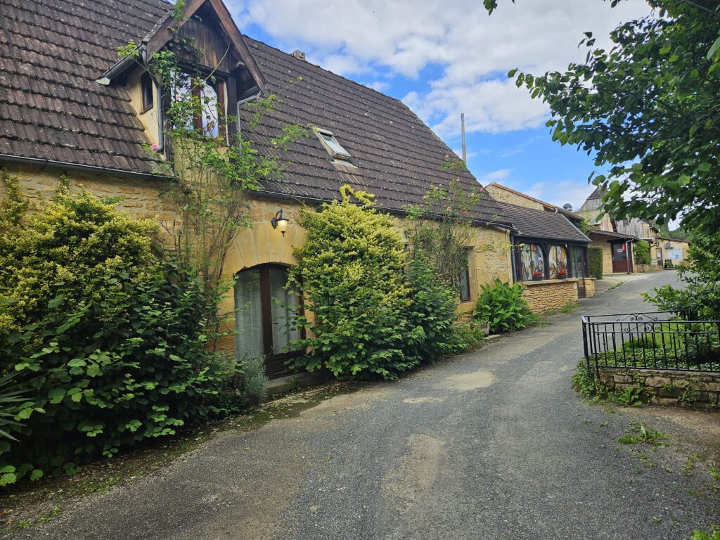 Charming traditional stone cottage at a French campsite in Dordogne, surrounded by lush greenery and a gravel path under a blue sky.