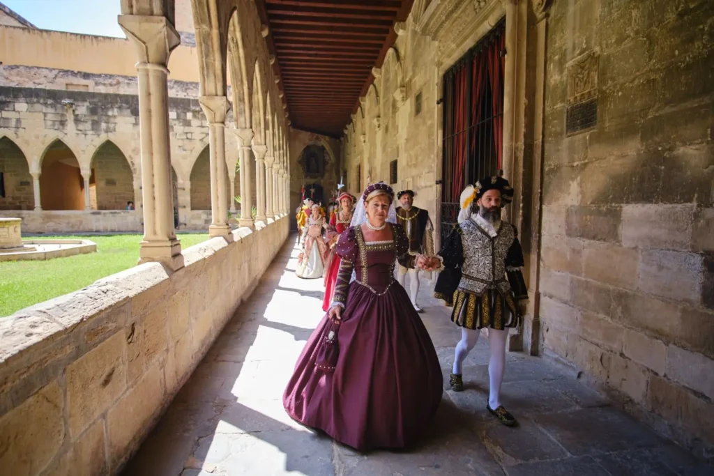 Pareja con vestimenta noble del siglo XVI caminando de la mano por un claustro de piedra con arcos, seguidos por una comitiva, durante la Festa del Renaixement de Tortosa.
