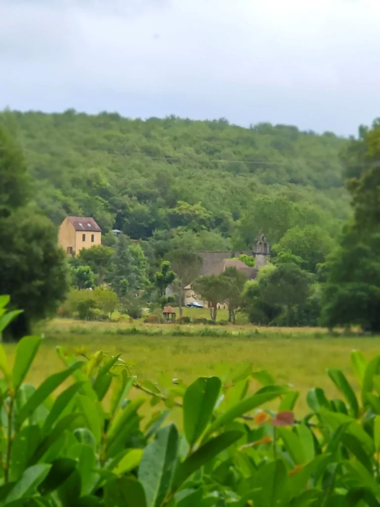 Vista lejana de una casa tradicional de piedra y la iglesia de Sant André de Allas rodeada de espeso bosque verde en el Périgord Noir, Francia. Paisaje rural representativo del slow travel y destinos sin masificar.