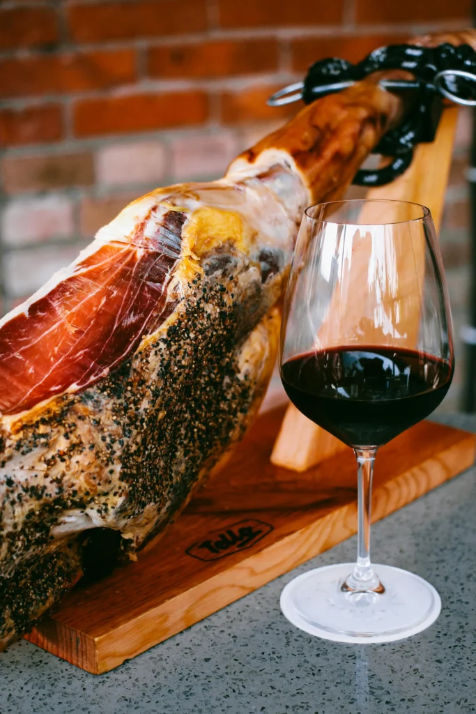 Jamón ibérico con una copa de vino tinto sobre soporte de madera, presentación gastronómica elegante y tradicional.