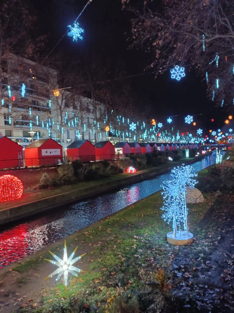 Casetas de madera rojas iluminadas junto a un canal en un mercadillo de Navidad tradicional en Francia, sin aglomeraciones.