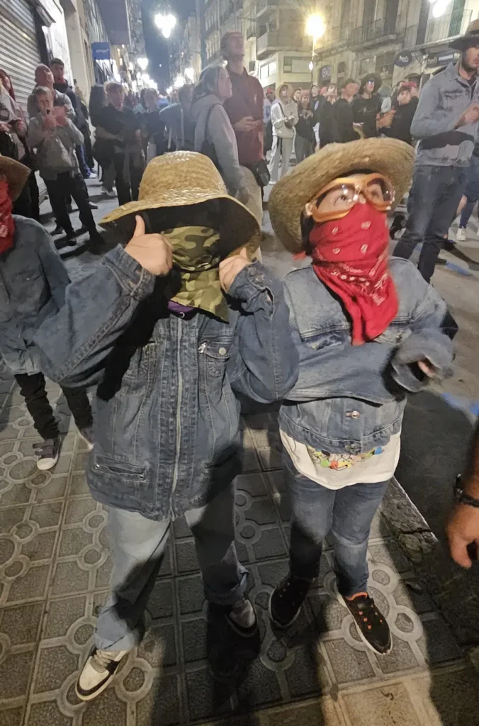 Dos niños, equipados para un Correfoc con chaquetas y pantalones vaqueros, sombreros de paja, pañuelos cubriendo la cara y gafas de protección.