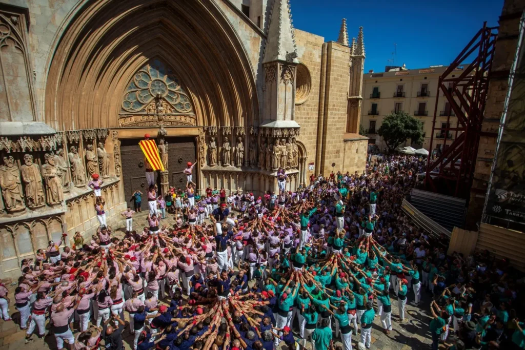 Multitud de personas con camisas de colores formando una torre humana (castell) frente a la fachada principal de la Catedral de Tarragona en un día soleado.