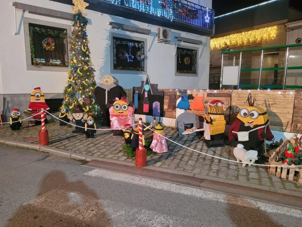 Decoración navideña callejera y artesanal con figuras hechas a mano tipo Minion disfrazadas alrededor de un árbol de Navidad en la acera de una calle de pueblo.