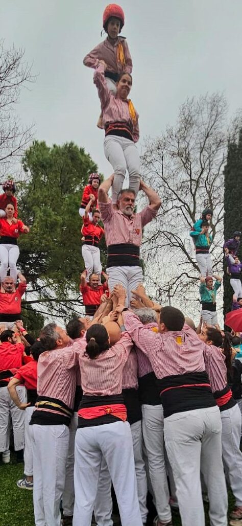 Un grupo de castellers practica una torre humana (castell) en un parque de hierba rodeado de árboles con follaje de otoño bajo un cielo gris. La joven con casco rojo está en la cima.