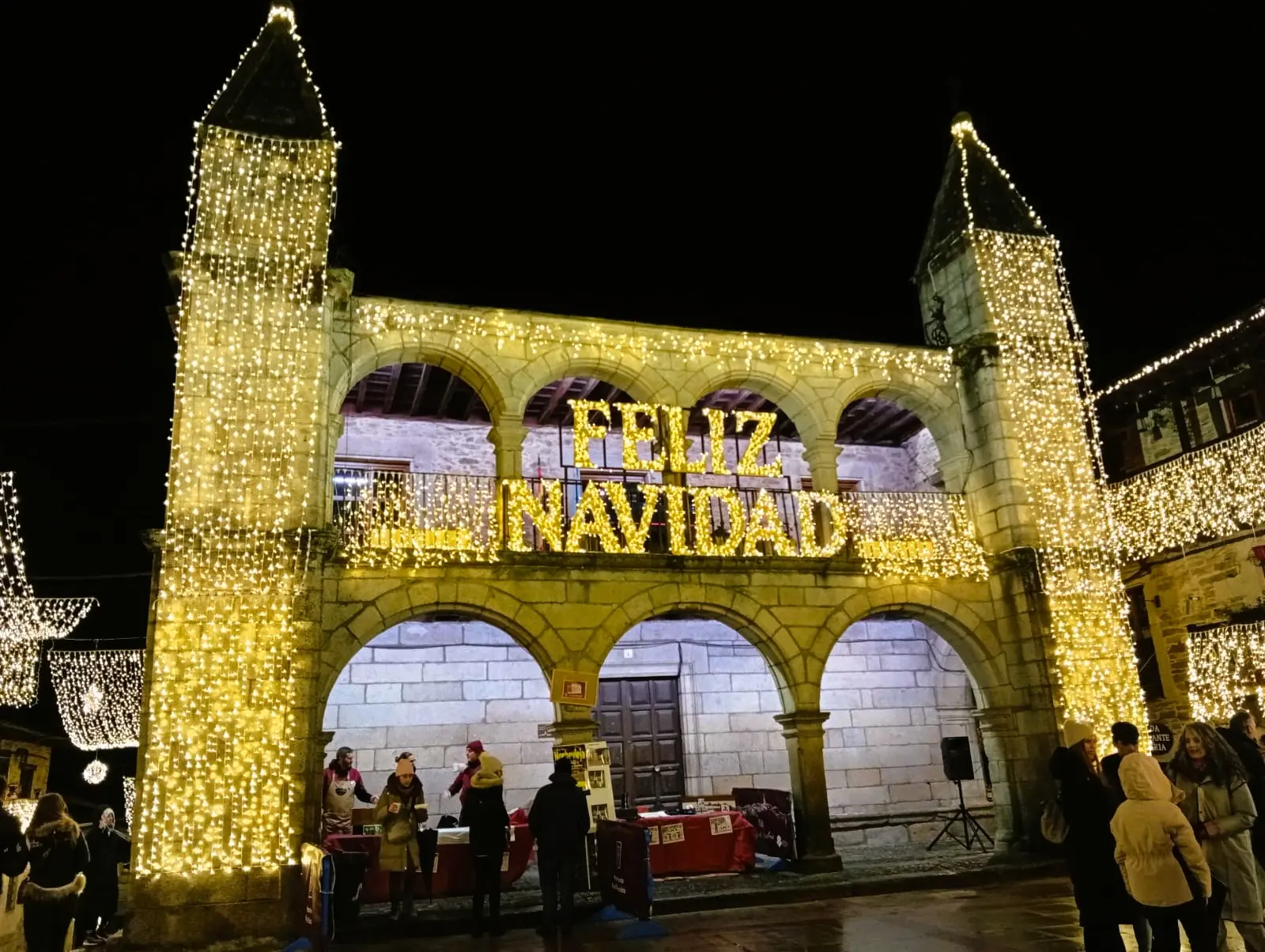 Fachada de piedra de un edificio histórico con arcos y dos torres, cubierta completamente de luces doradas formando las palabras Feliz Navidad, con personas paseando debajo.