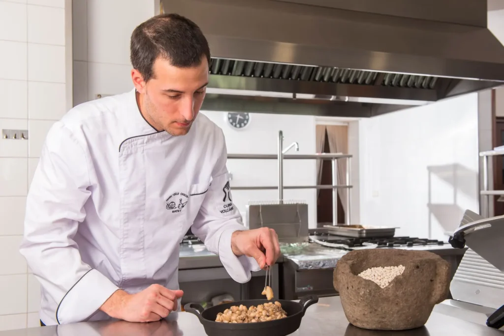 Chef con chaquetilla de la Cuina Volcànica emplatando cuidadosamente unas alubias tradicionales junto a un mortero de piedra rústica en una cocina profesional.