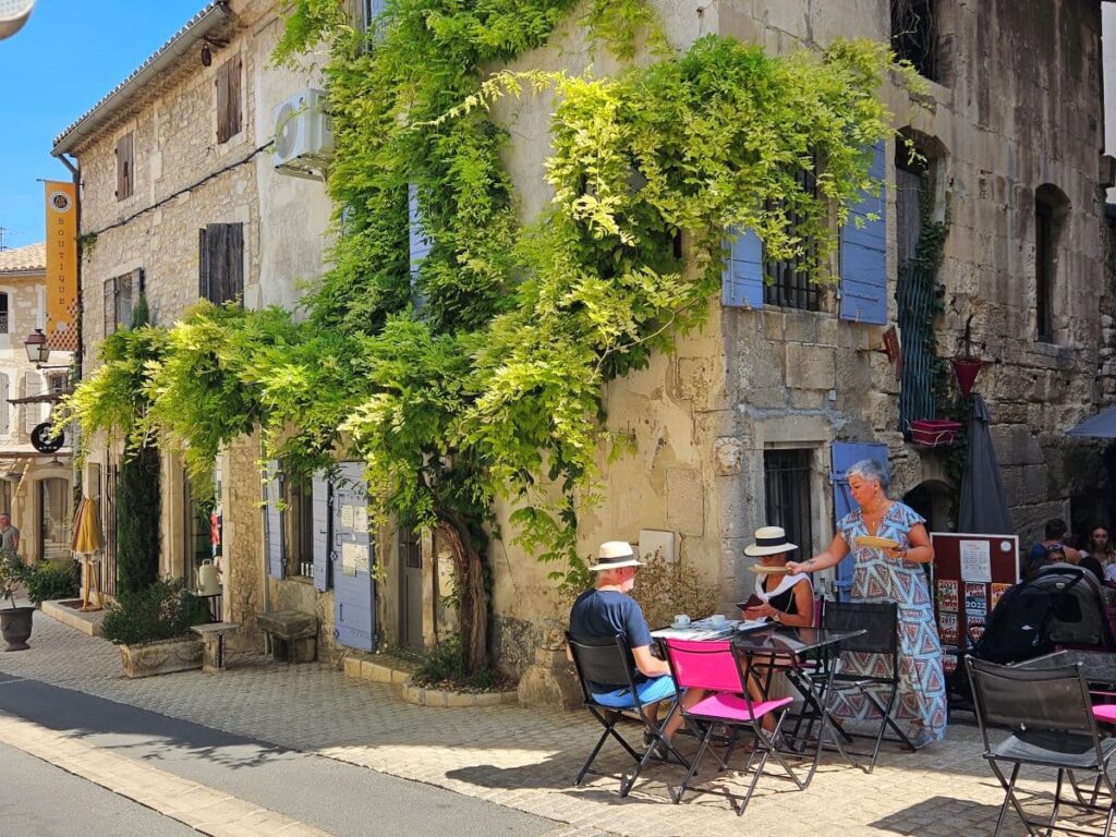Pareja comiendo en una pequeña terraza al aire libre junto a un edificio histórico de piedra con contraventanas azules y una gran enredadera en la Provenza.