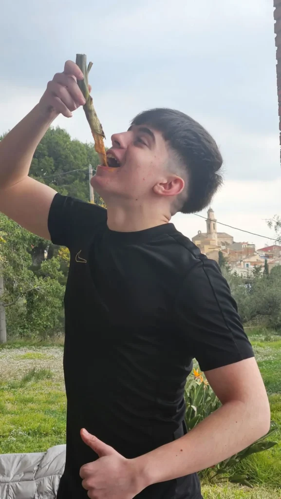 Chico joven comiendo un calçot untado en salsa romesco al aire libre en Botarell, con el campanario del pueblo de fondo.