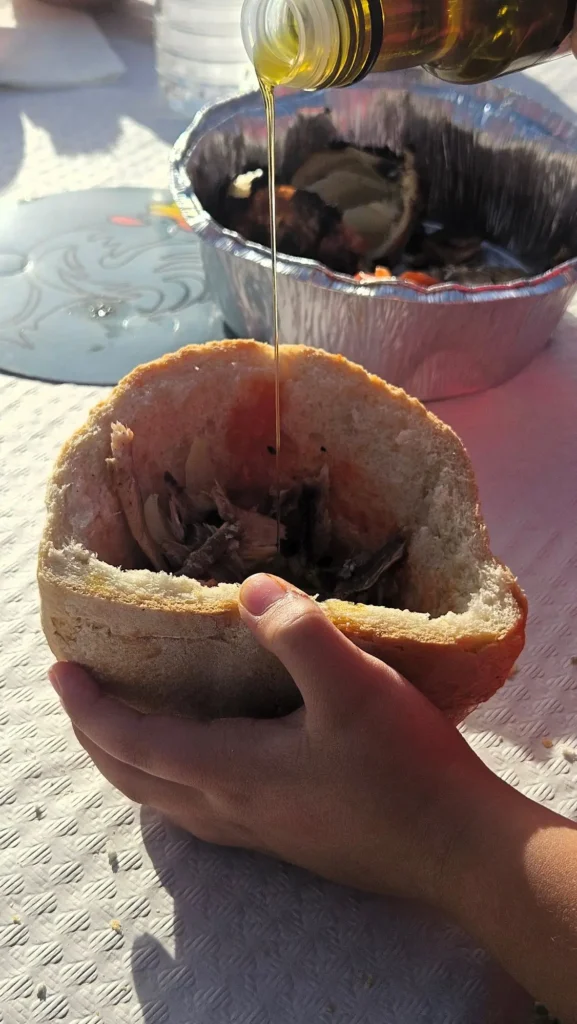 Un chorro de aceite de oliva virgen extra cayendo sobre el interior de un pan de payés vaciado y relleno, sostenido por unas manos al sol.