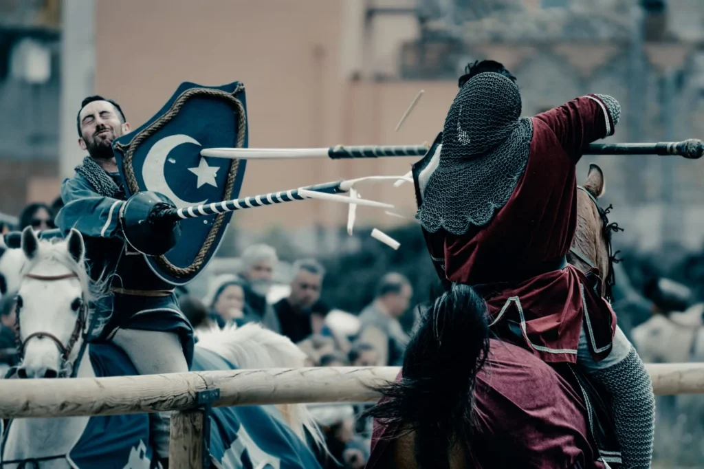 Una foto de acción de dos caballeros medievales a caballo en una barrera de madera durante un torneo de justas. Sus lanzas de madera se están rompiendo y astillando en el impacto. El caballero de la izquierda lleva un escudo azul con una luna creciente y una estrella blancas. El caballero de la derecha, de espaldas, lleva una cota de malla y una túnica roja. Un público borroso y edificios antiguos están en el fondo.