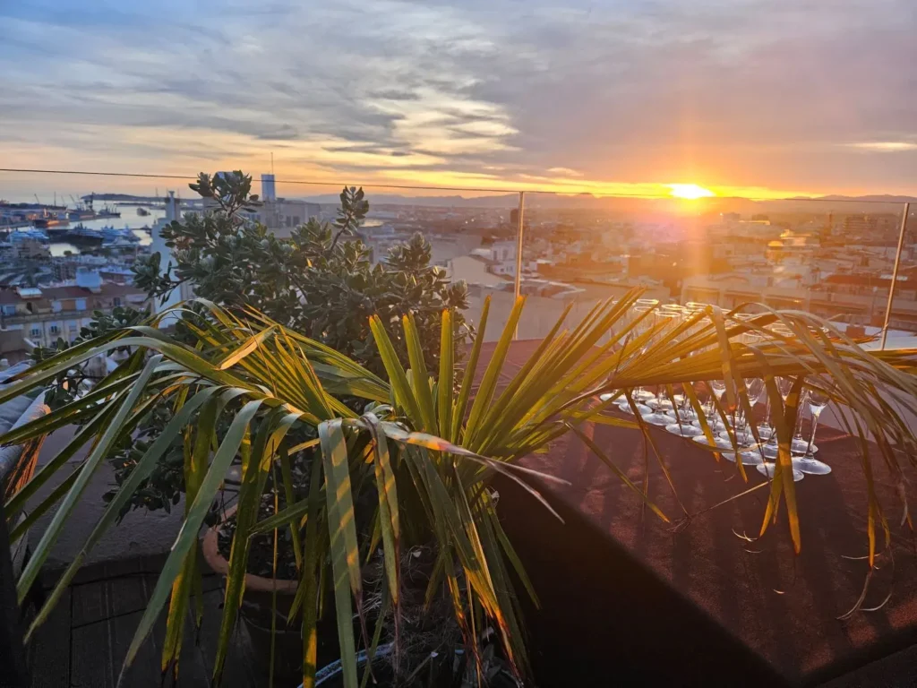 Vista panorámica de un atardecer dorado sobre la ciudad y el puerto de Tarragona desde una terraza con plantas y copas de cristal.